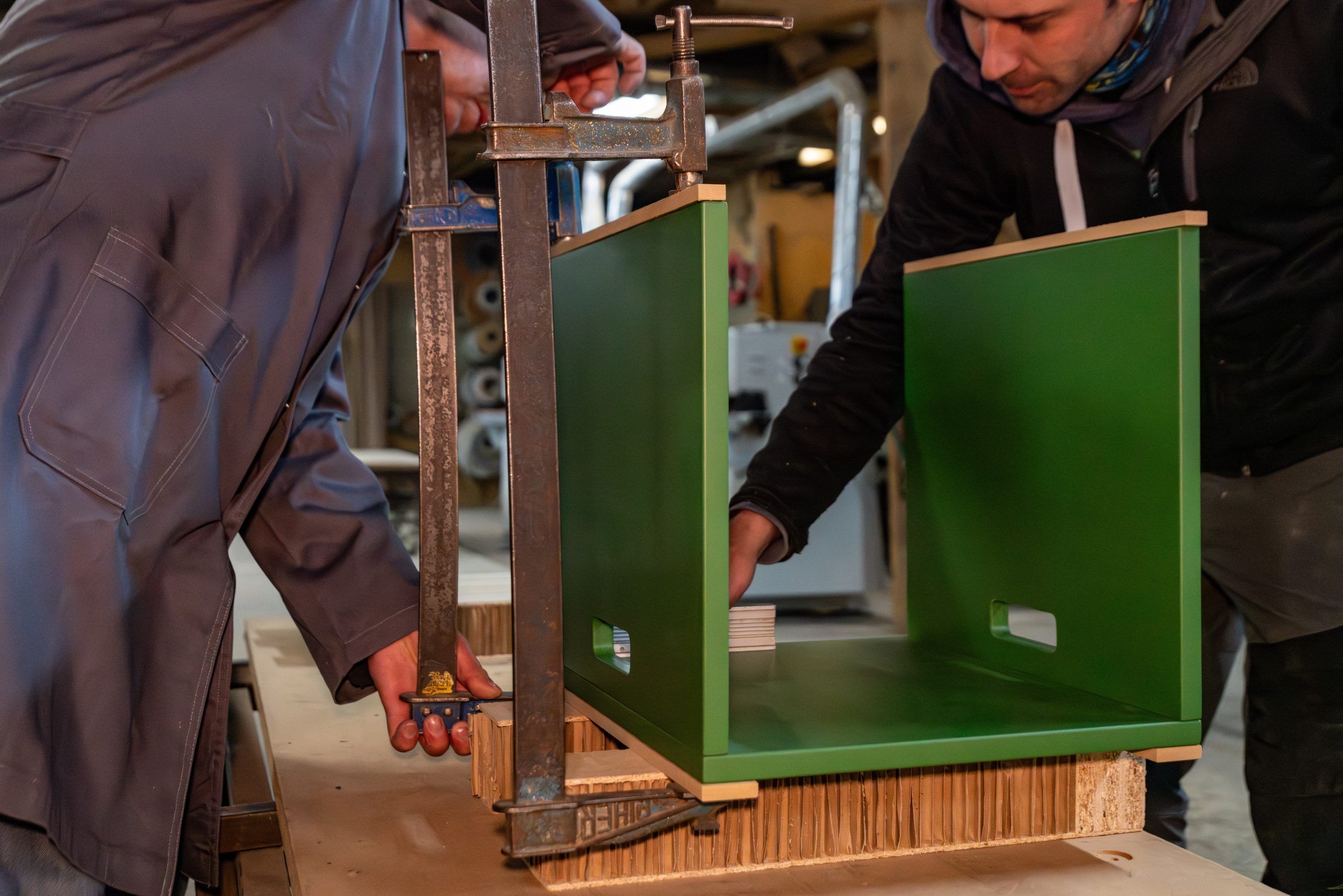 Dos artesanos de Madecsa Atelier trabajando en el ensamblaje de una mesa lacada verde utilizando sargentos en su taller de Andorra.